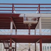 Worker on a suspended platform applying fire protection coating to red structural steel beams