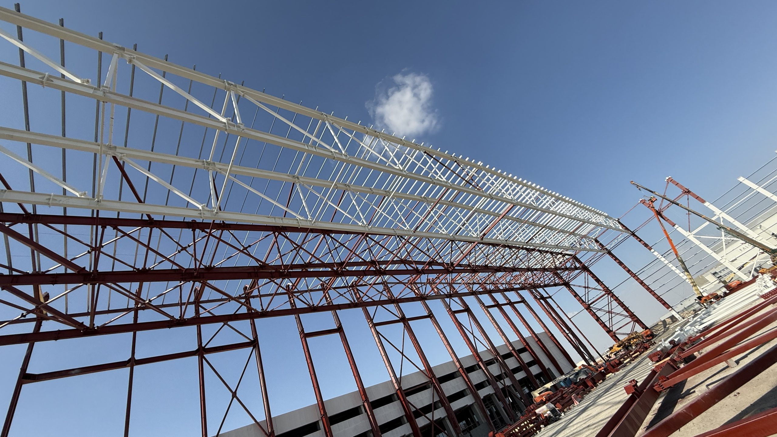 Wide-angle view of a red and white steel warehouse frame under construction with a crane